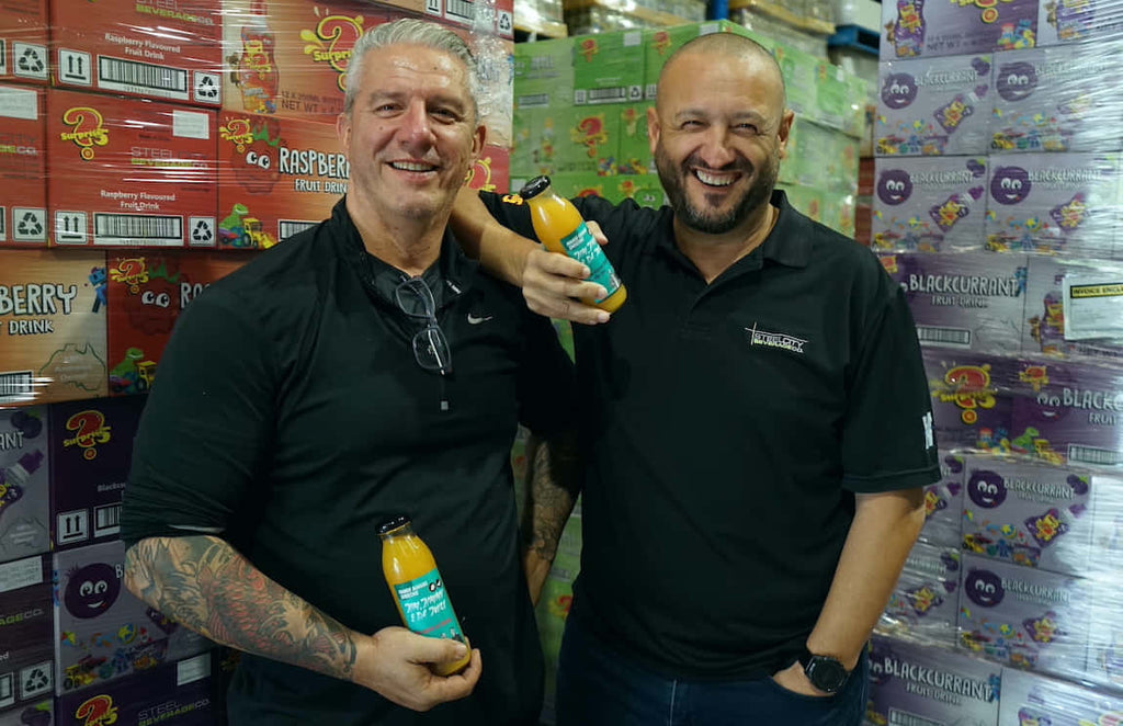 Two men holding juice bottles in their hands in front of the boxes with beverages in the warehouse